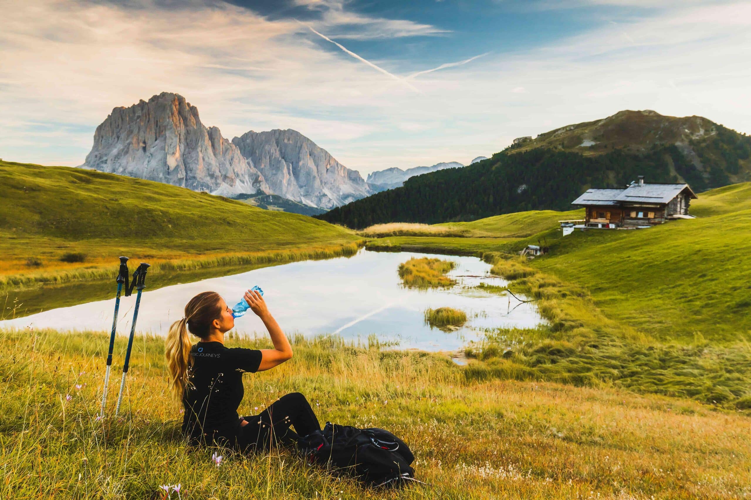 Woman drinking from a water bottle, taking a rest in front of a pond with Dolomites in the background, Italy.