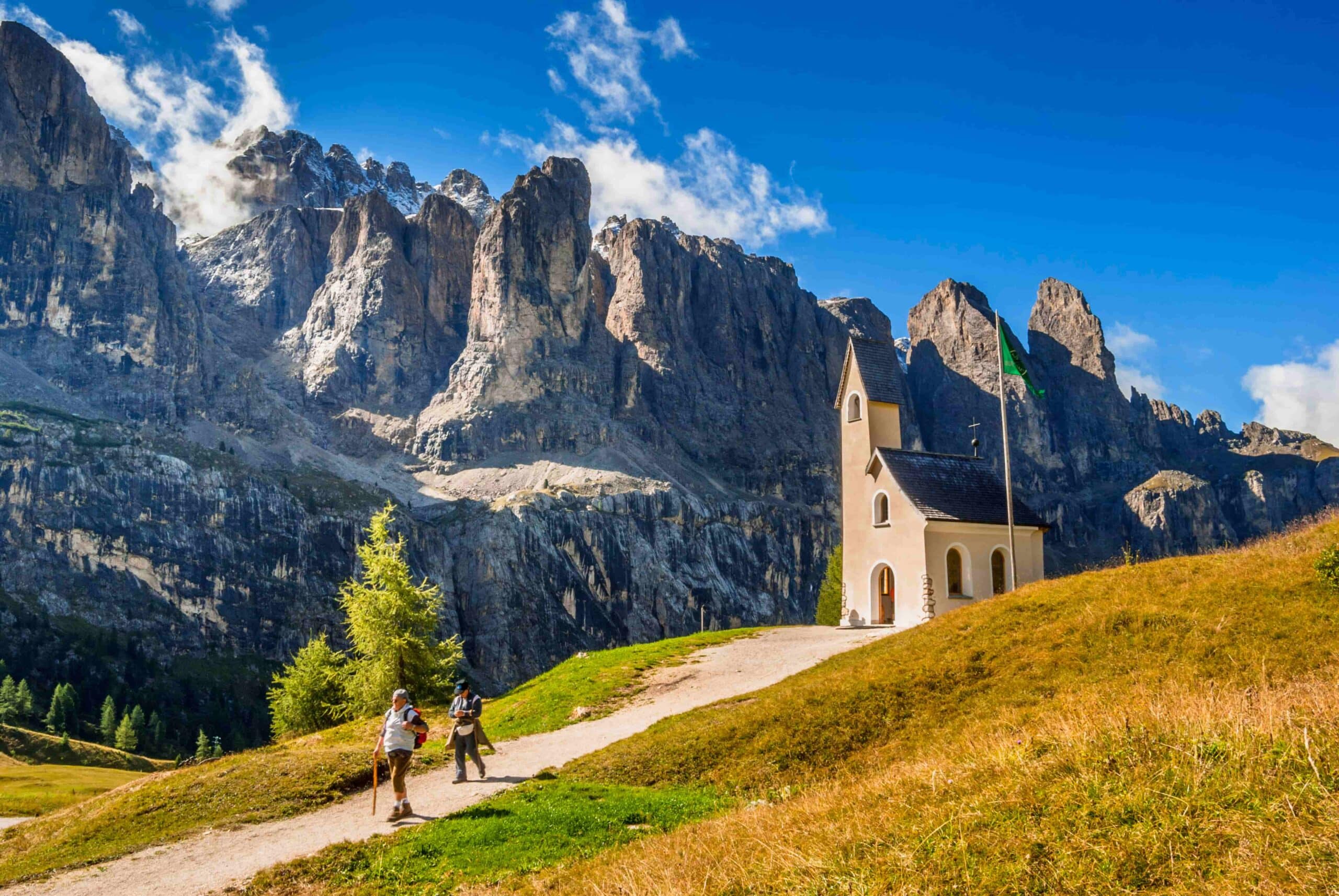 Two hhikers walk past a quaint church with Dolomite Mountain range in the background.